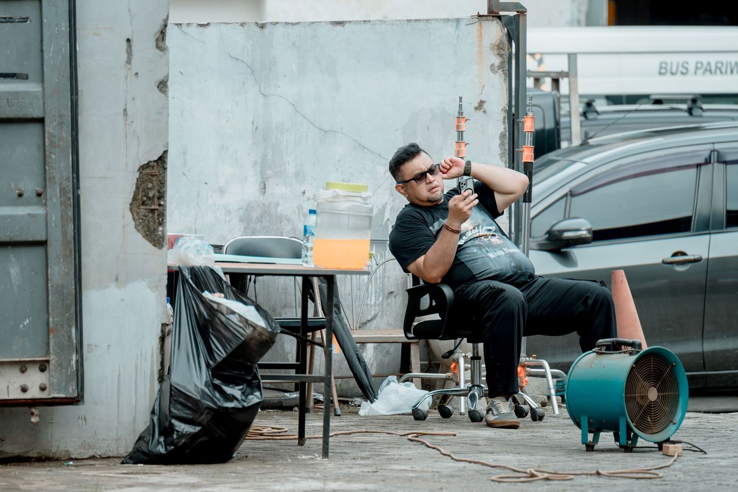 Man sitting outdoors near trash bags and a fan, holding a camera in an urban alleyway setting.