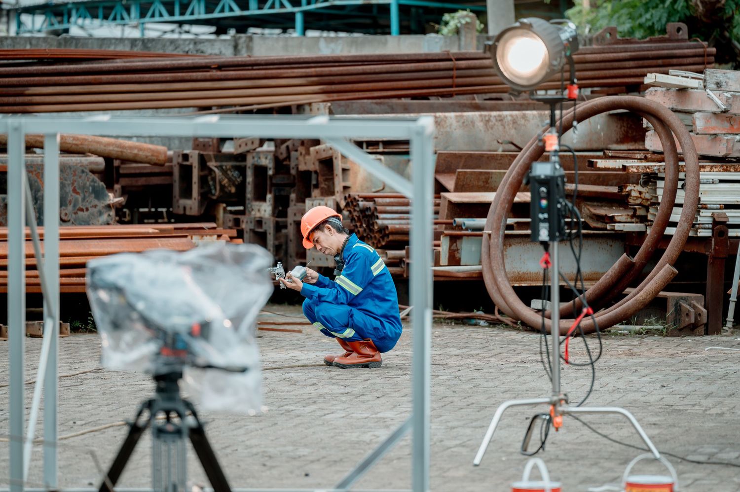 Worker in orange safety gear inspects metal pipes at an industrial site with professional lighting equipment nearby.