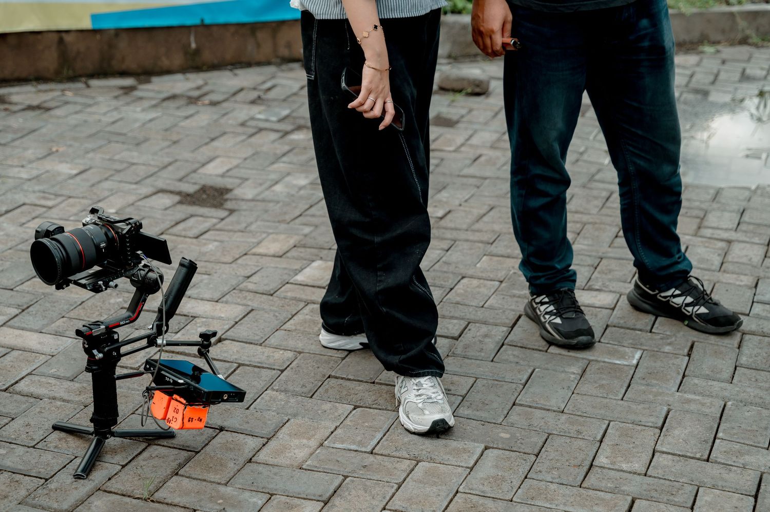 Two people standing on brick pavement next to a professional camera gimbal stabilizer setup outdoors.