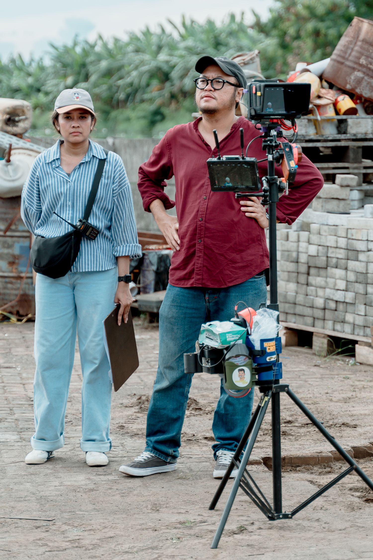 Two filmmakers stand beside a camera on tripod at an outdoor film set with palm trees in background.