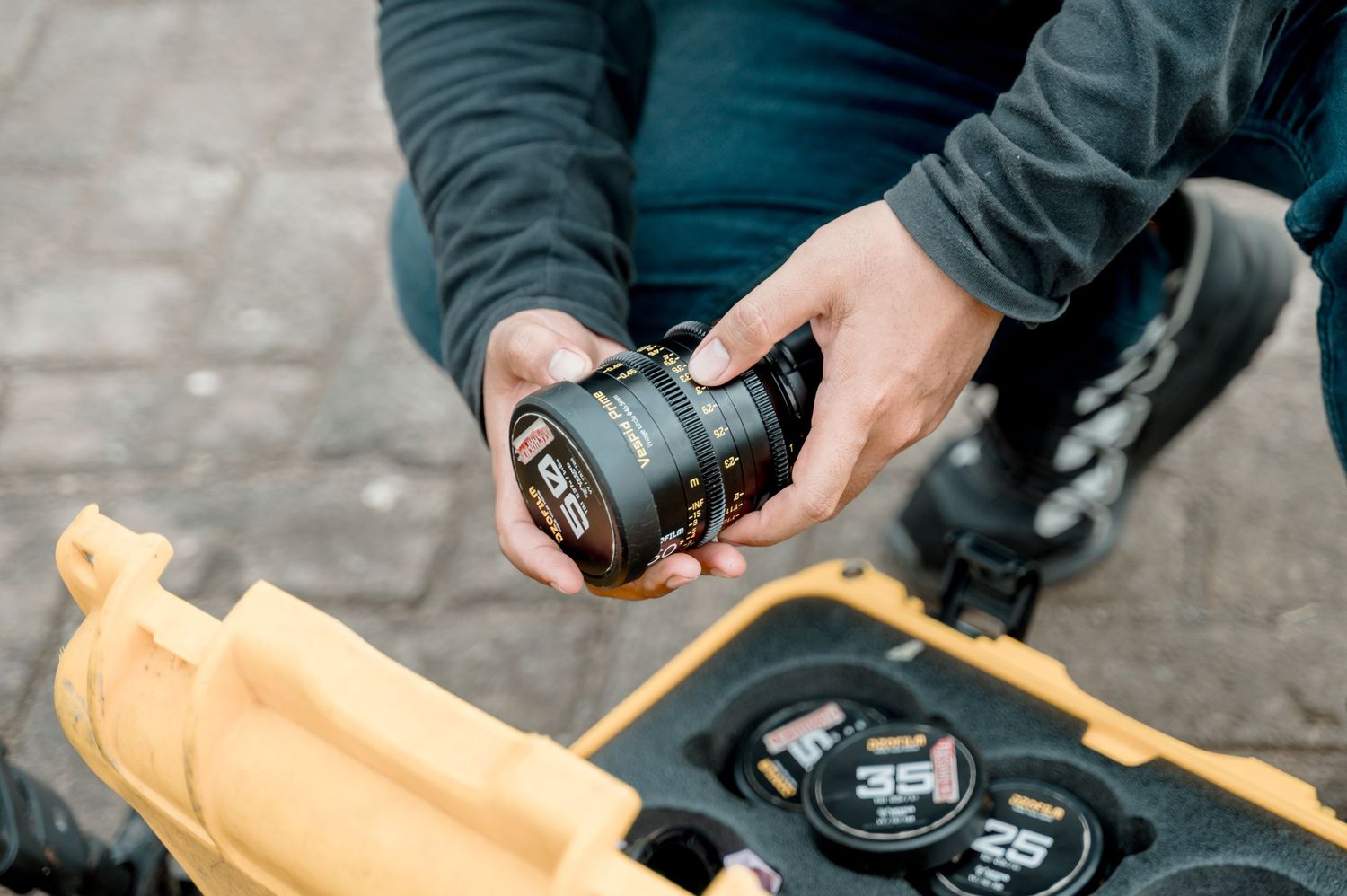 Person holding a cinema camera lens over a yellow Pelican case with multiple lenses on a film set.
