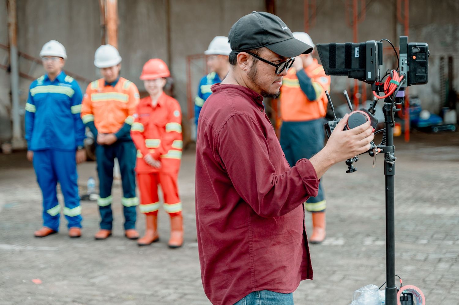 A filmmaker adjusts a camera on a tripod at an industrial site with workers in safety gear standing behind him.