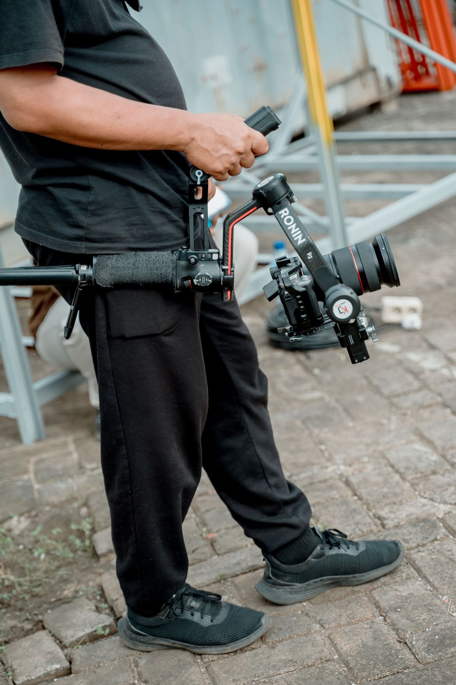 Videographer holding a professional camera gimbal stabilizer outdoors on a cobblestone surface.