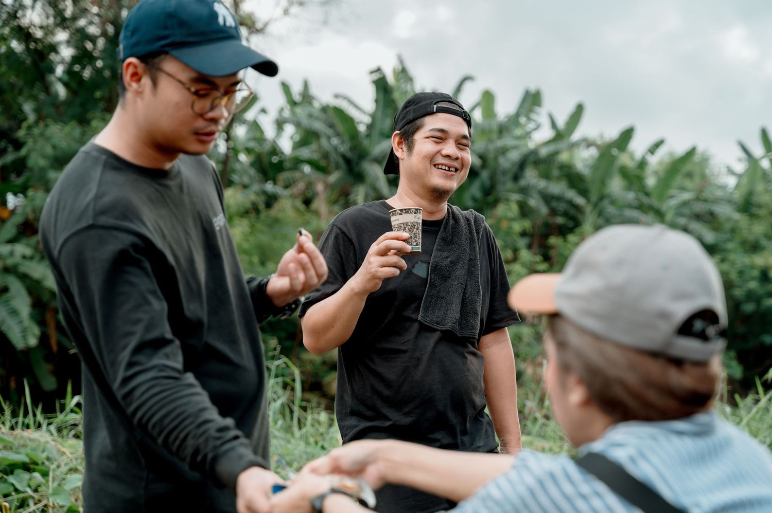 Three young men in black outfits socializing outdoors in a lush green tropical garden setting.