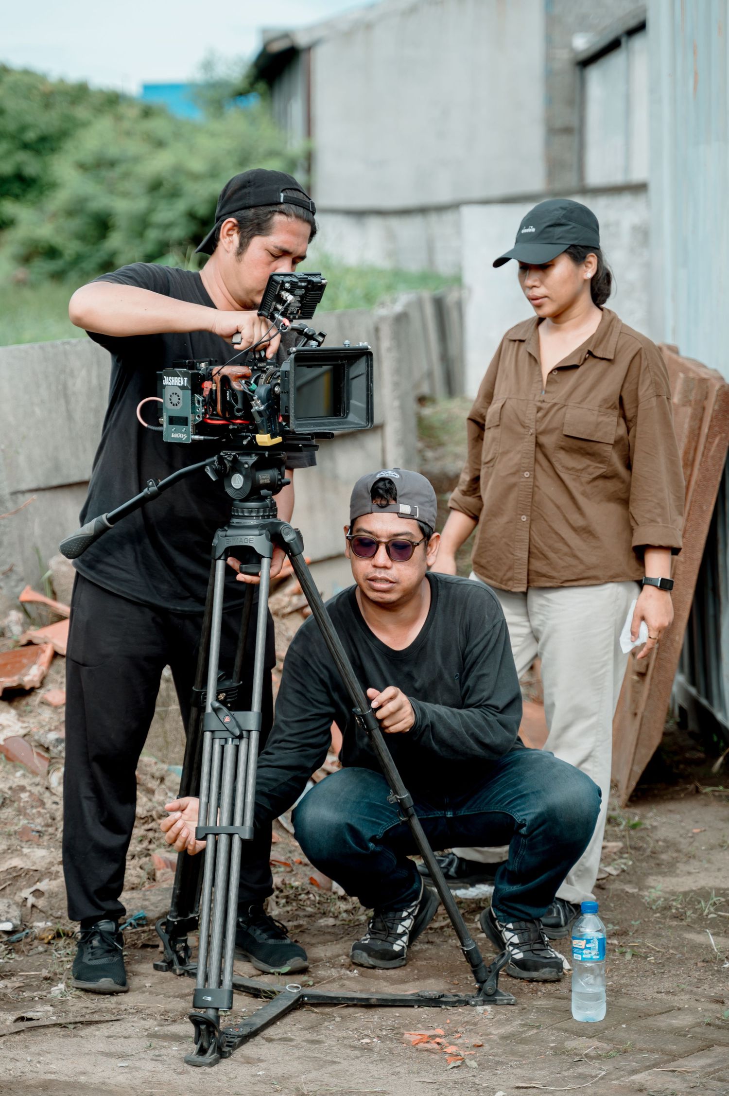 Film crew with camera on tripod shooting on location outdoors, two men and a woman collaborating on video production.