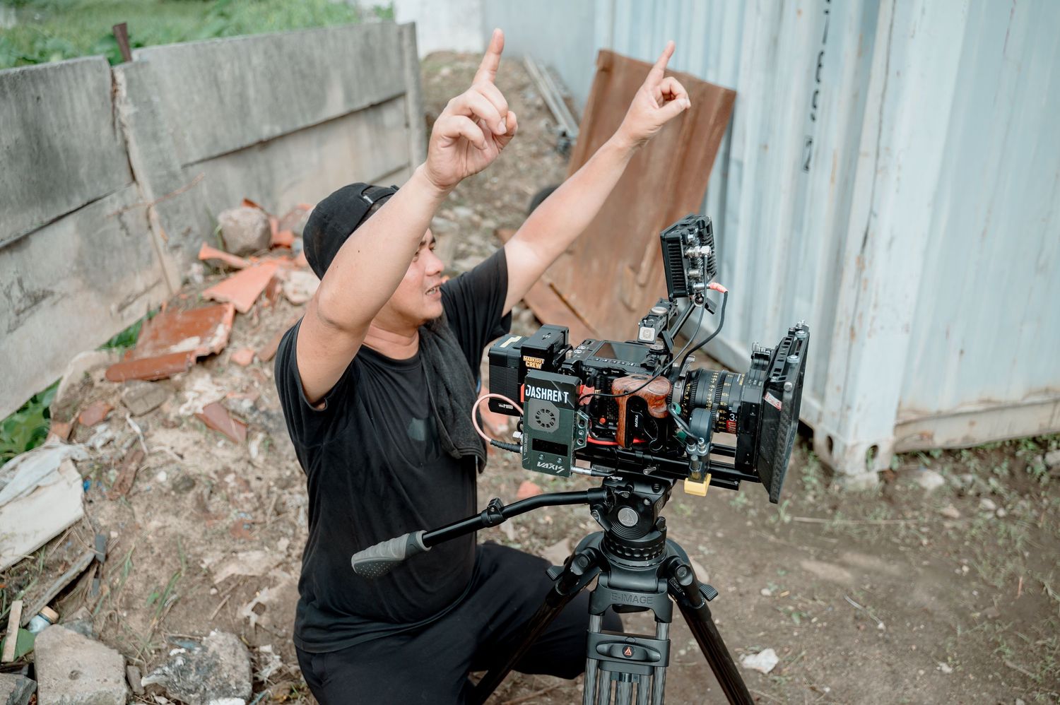 Filmmaker kneeling outdoors pointing up while operating a professional video camera on a tripod at a film shoot location.