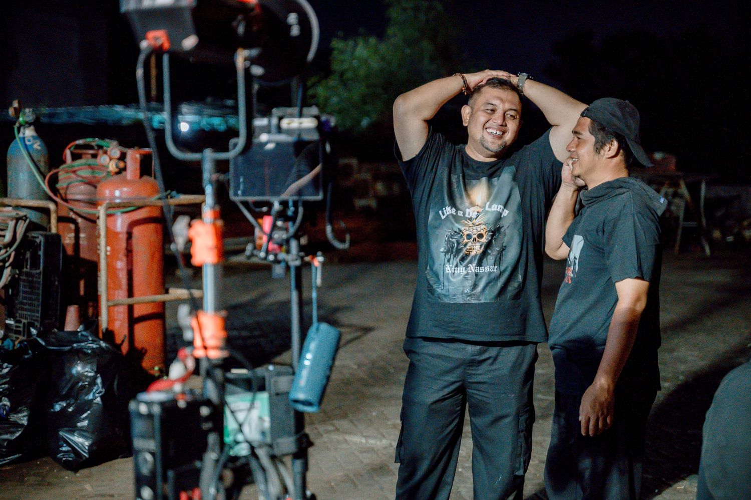 Two men laughing on a film set at night, surrounded by camera equipment and orange gas cylinders.