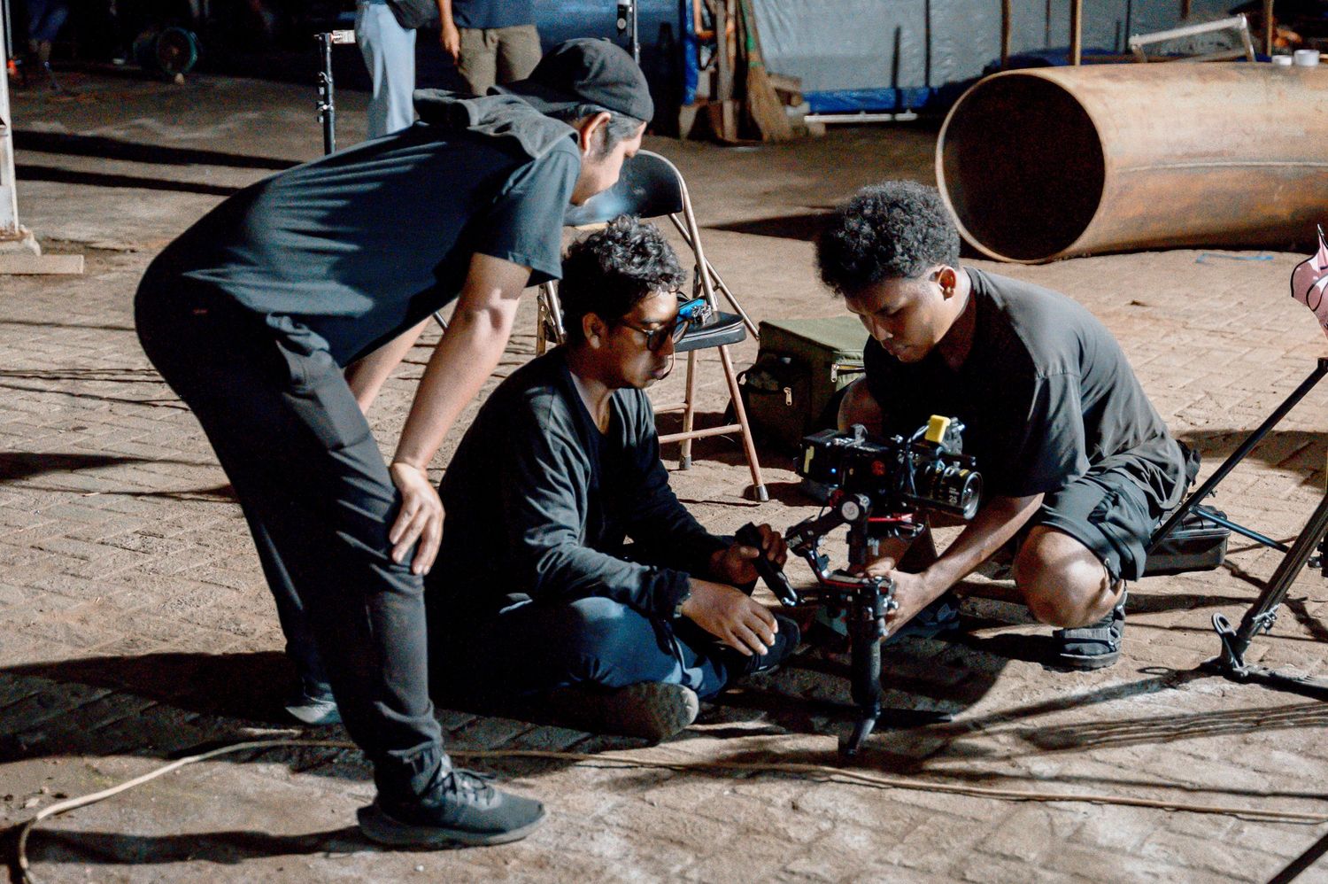 Three filmmakers collaborate on a camera setup on an industrial film set with large pipes in the background.