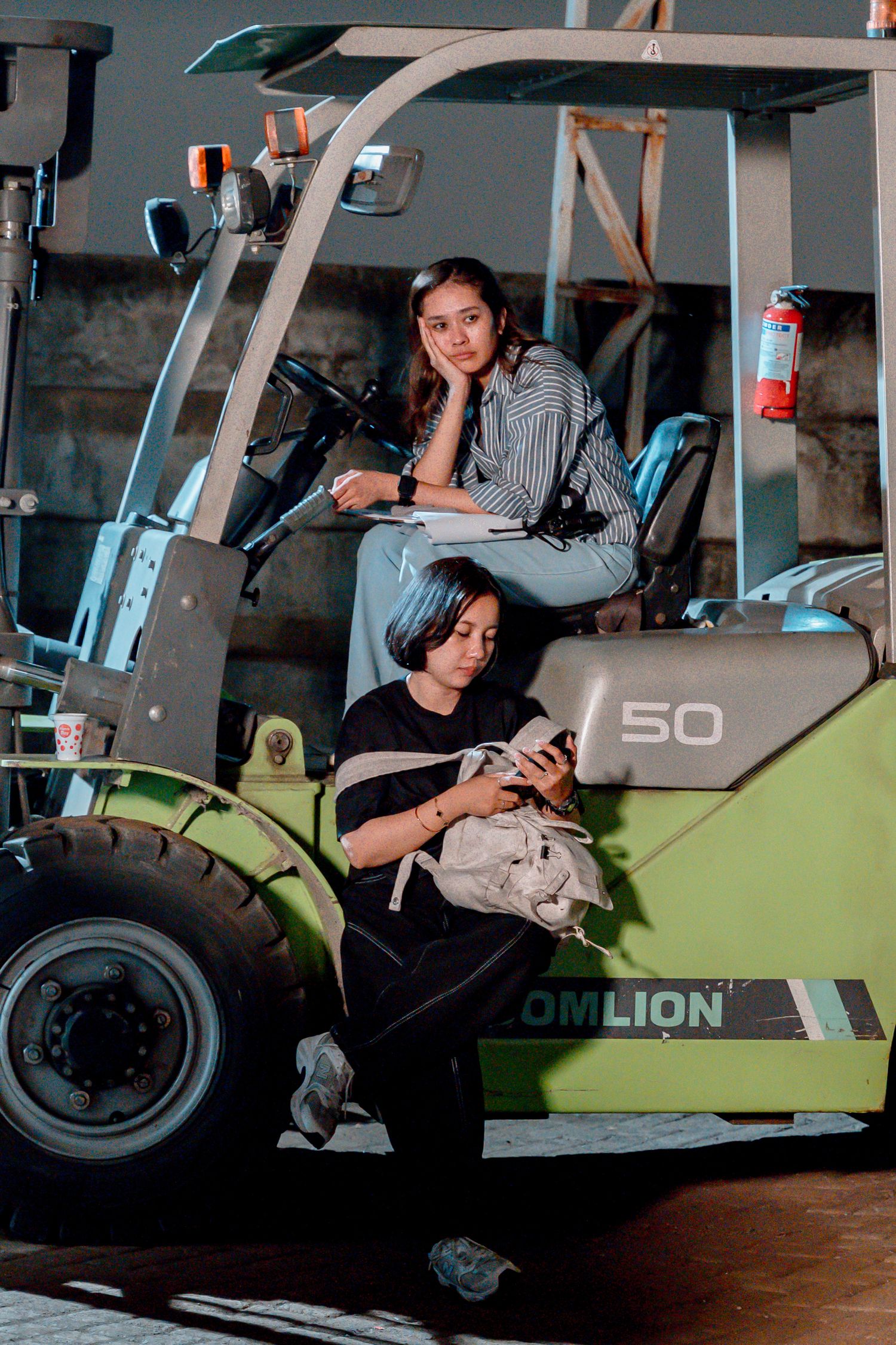 Two women posing near a green XCMG LOMLION forklift, one sitting on it and one leaning against it holding a phone.