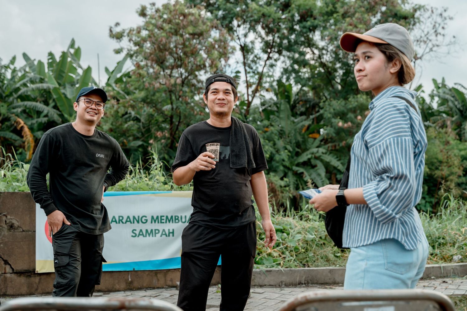Three people stand outdoors near a sign reading 'Jangan Membuang Sampah' surrounded by lush greenery.