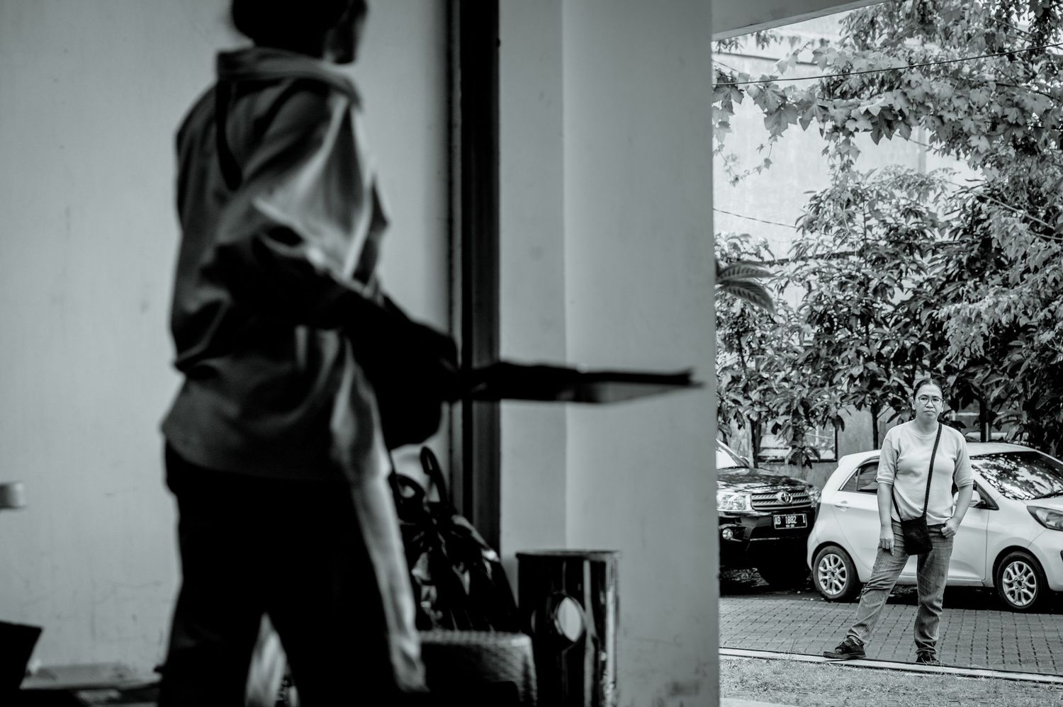 Black and white photo of a person pointing outside a doorway toward a man walking near parked cars on a tree-lined street.