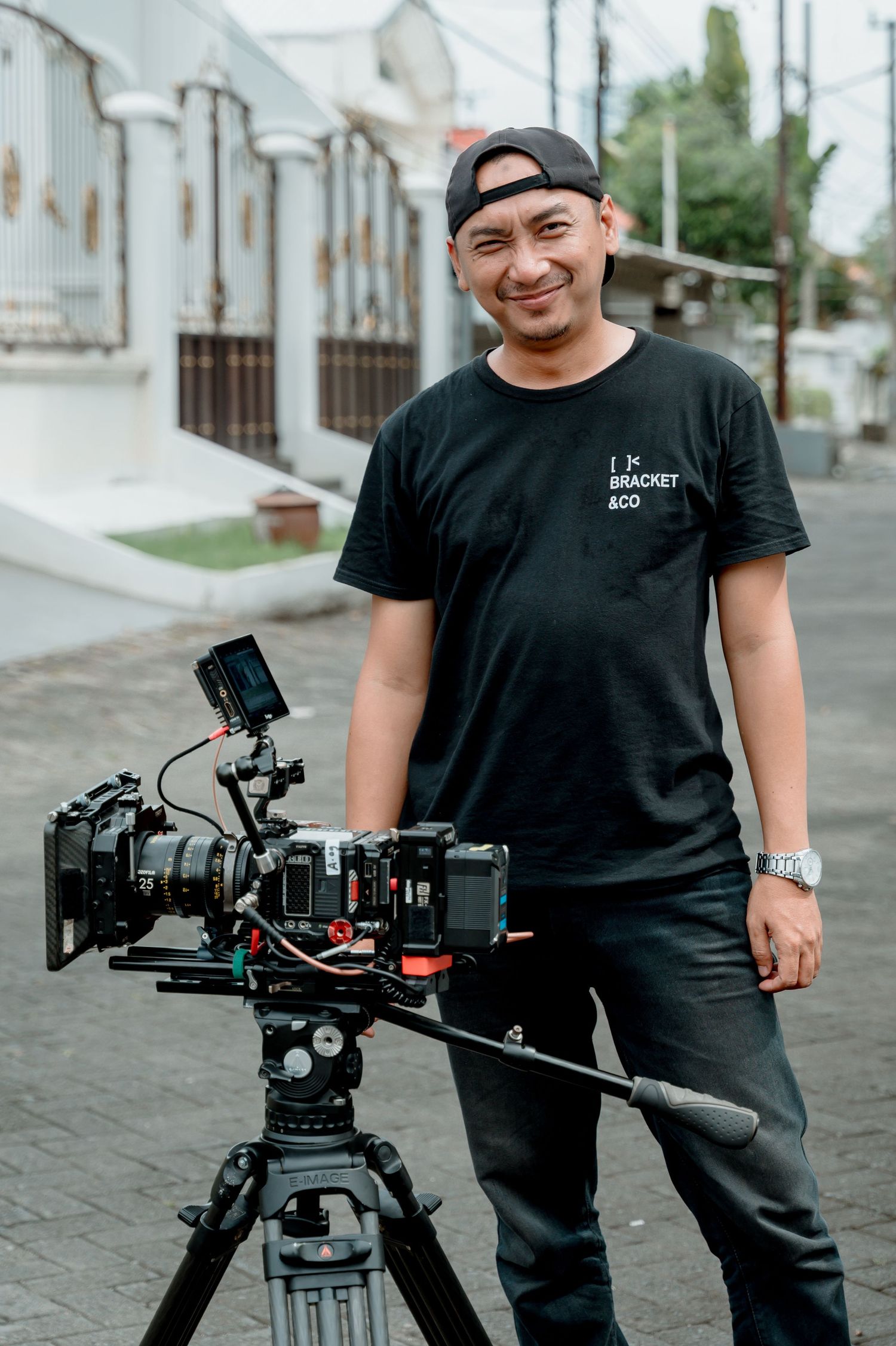 Smiling videographer in black t-shirt and cap standing beside professional cinema camera on tripod outdoors.