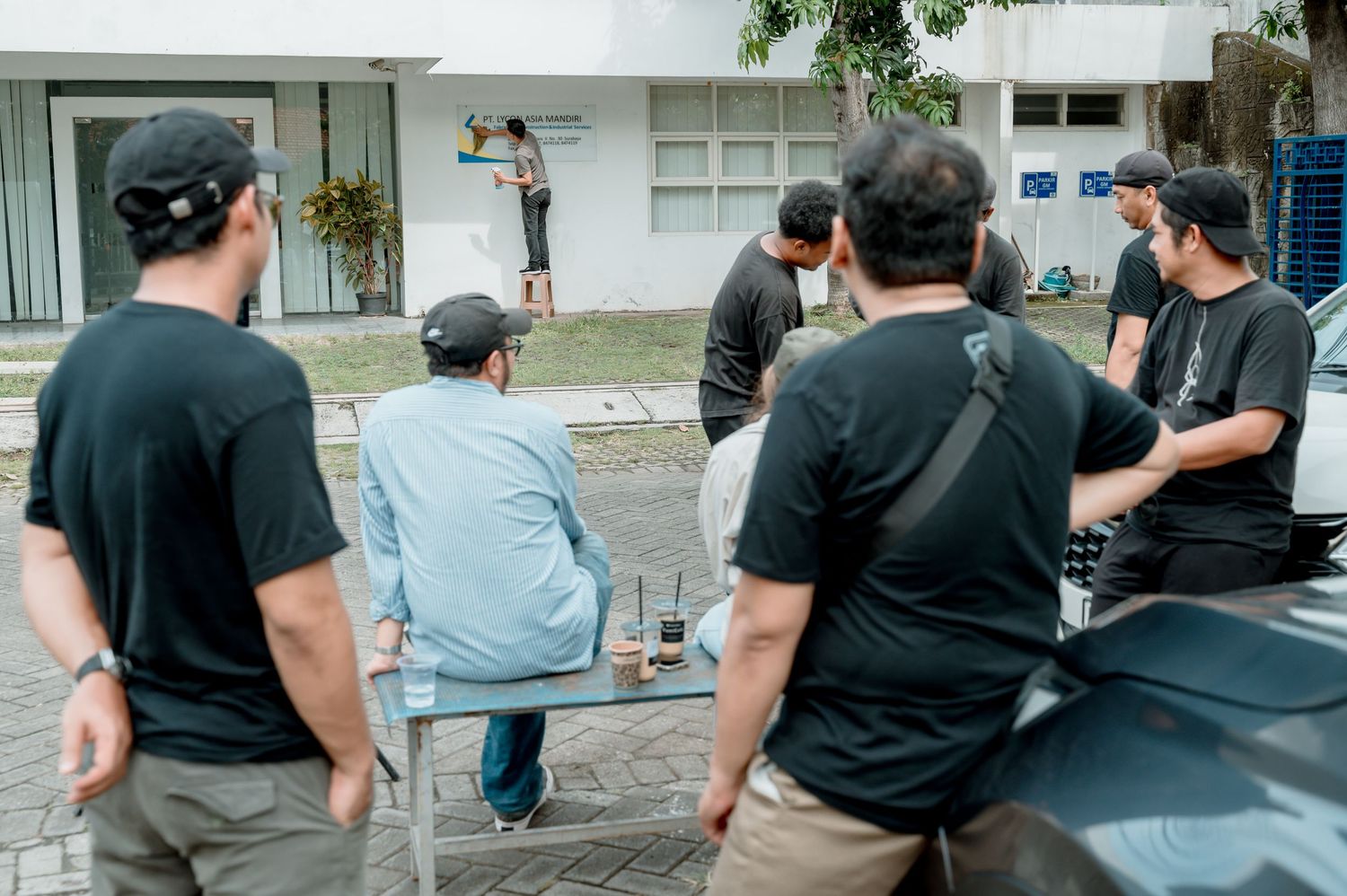 Group of people in black shirts watching an artist paint a mural on a white building outdoors.