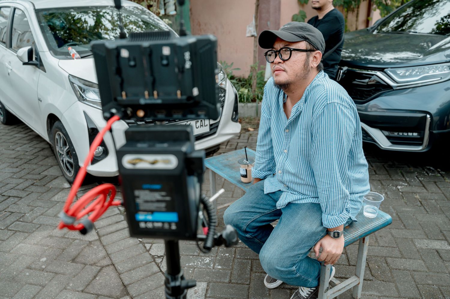 A film director wearing glasses and a cap sits on a stool reviewing a shot on a professional camera monitor.