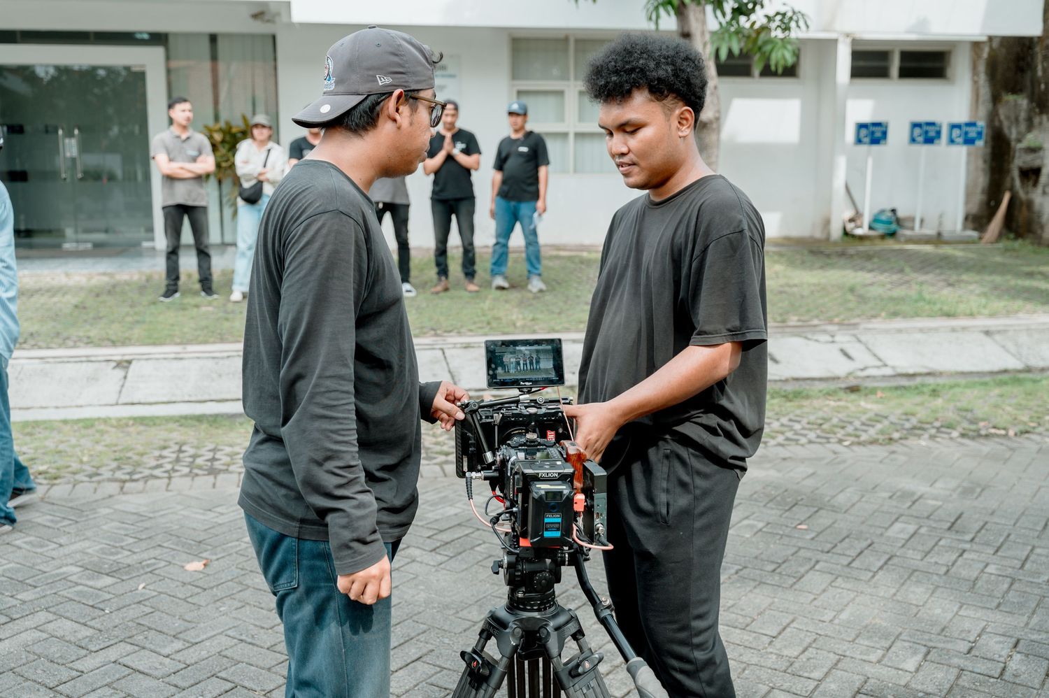 Two filmmakers in black shirts operating a professional cinema camera on a tripod outdoors on a film set.