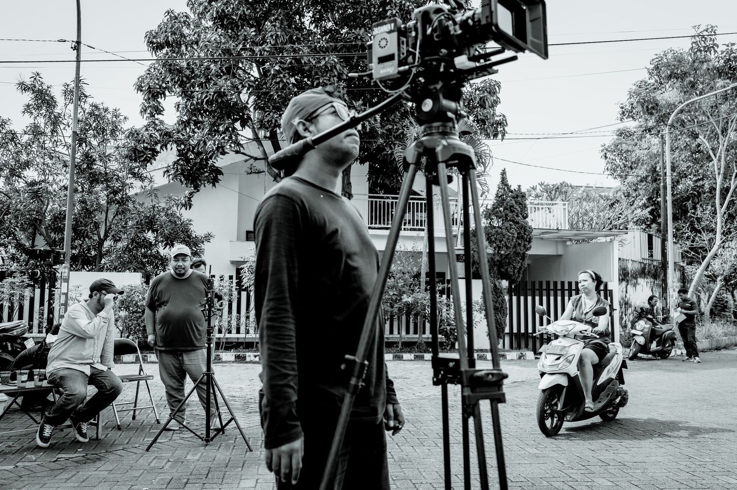 Black and white photo of a filmmaker operating a film camera on a tripod outdoors on a film set.