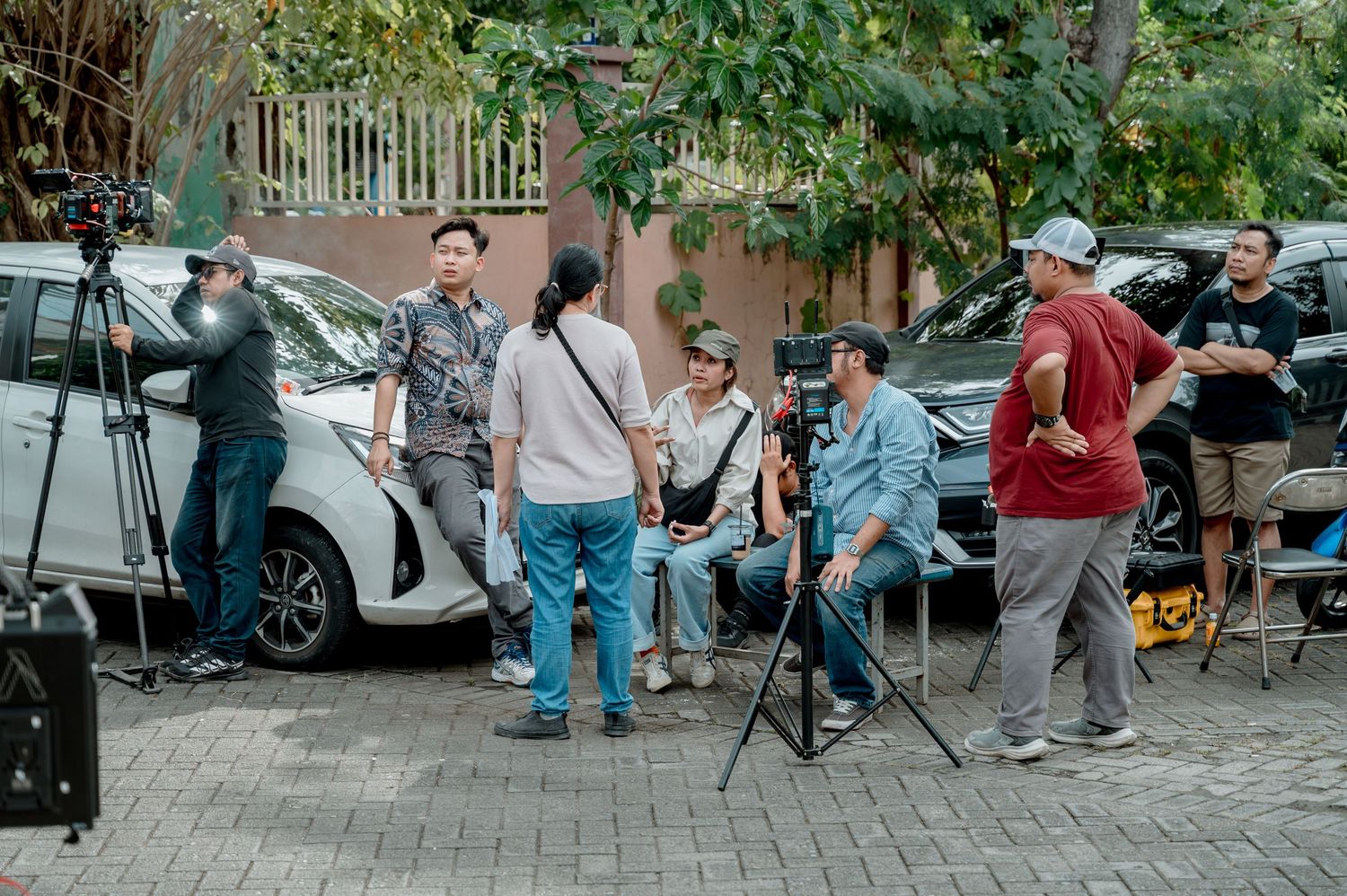 A film crew shoots an outdoor scene on a brick plaza with cameras and equipment surrounded by parked cars and trees.