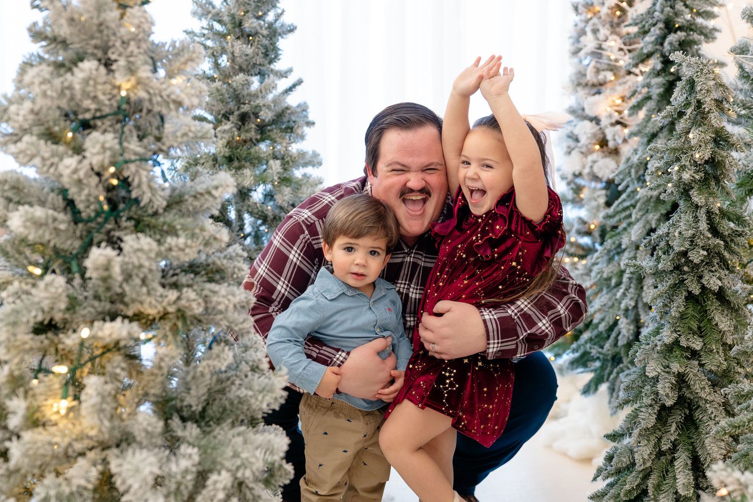 A cheerful father holds two young children among snow-flocked Christmas trees in a festive holiday family portrait.