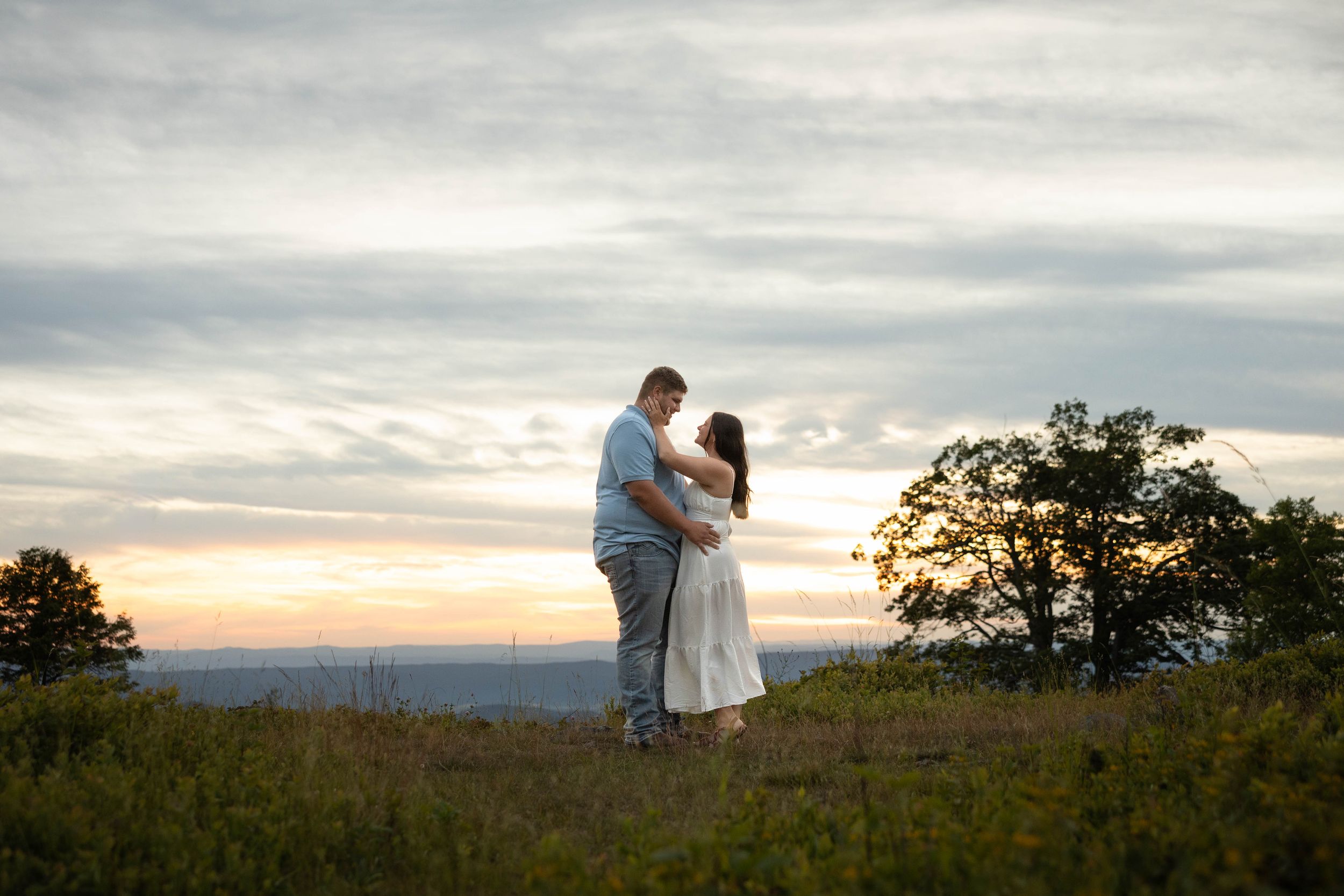 Couple embracing at sunset in a green field, woman smiling over man's shoulder, engagement ring visible.