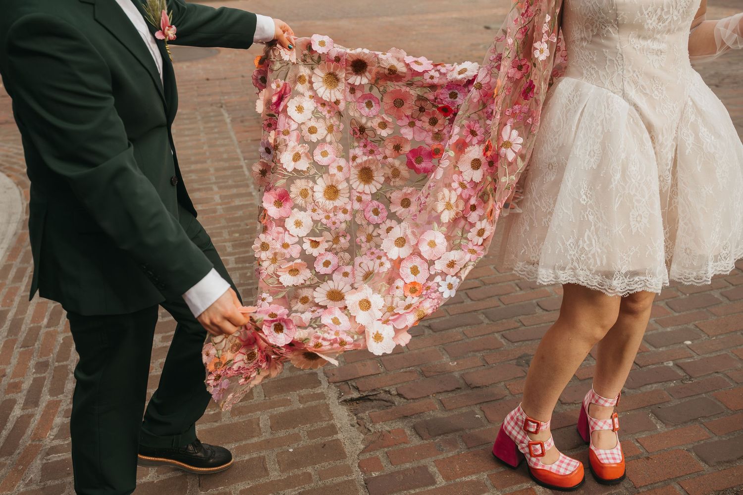 Bride and groom holding a floral pink cape with daisy print, bride wearing red platform shoes at a brick venue.