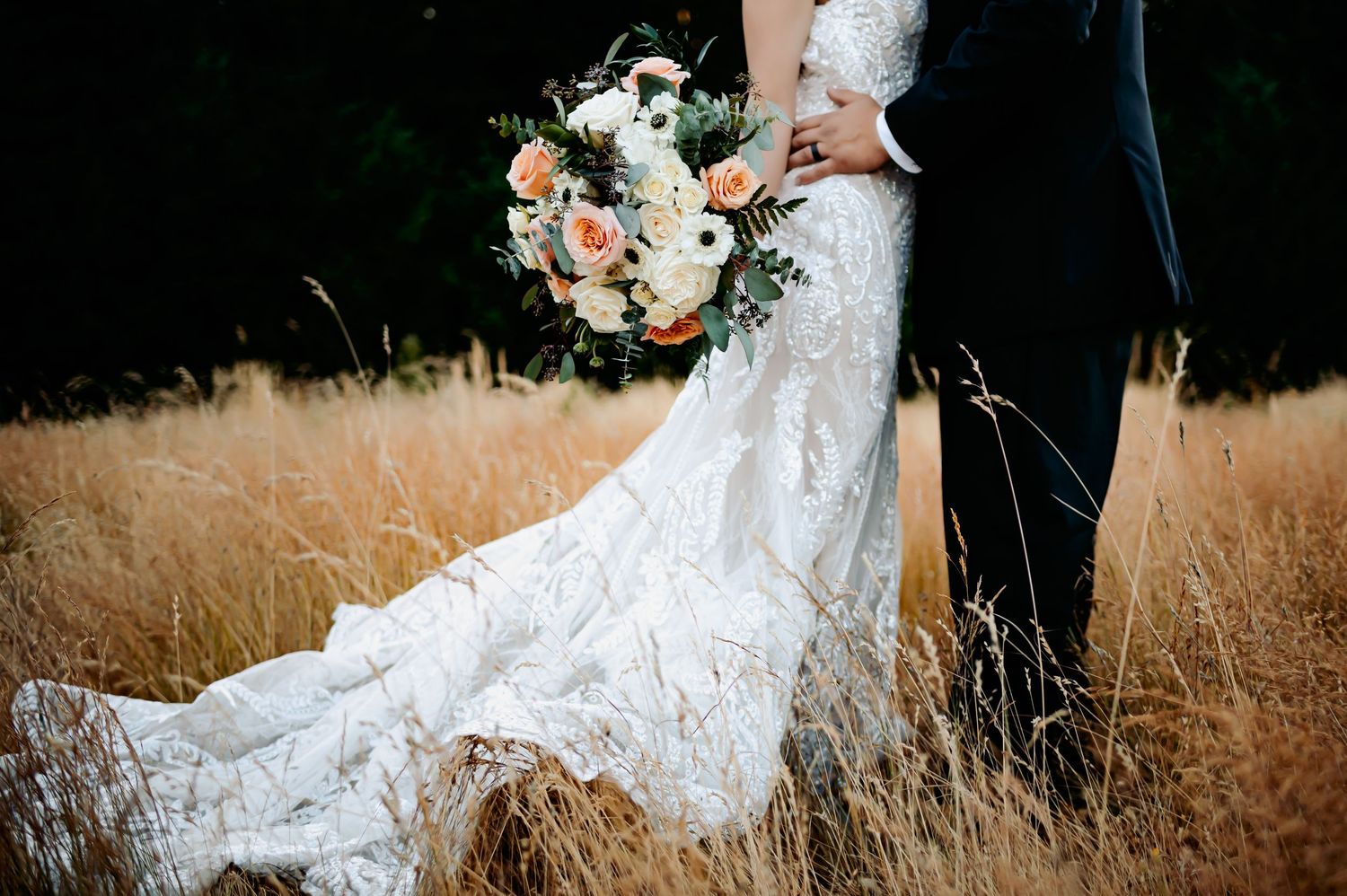 Bride in lace wedding gown with floral bouquet standing with groom in golden field.