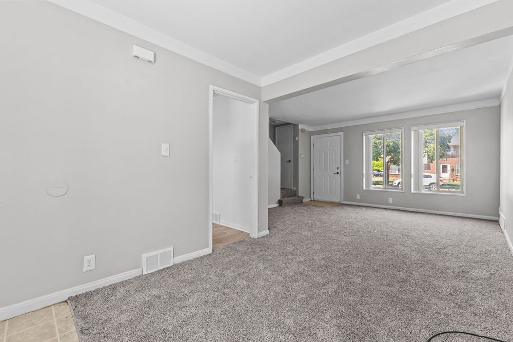Open living room with gray carpet, crown molding, white walls, and a window with natural light.