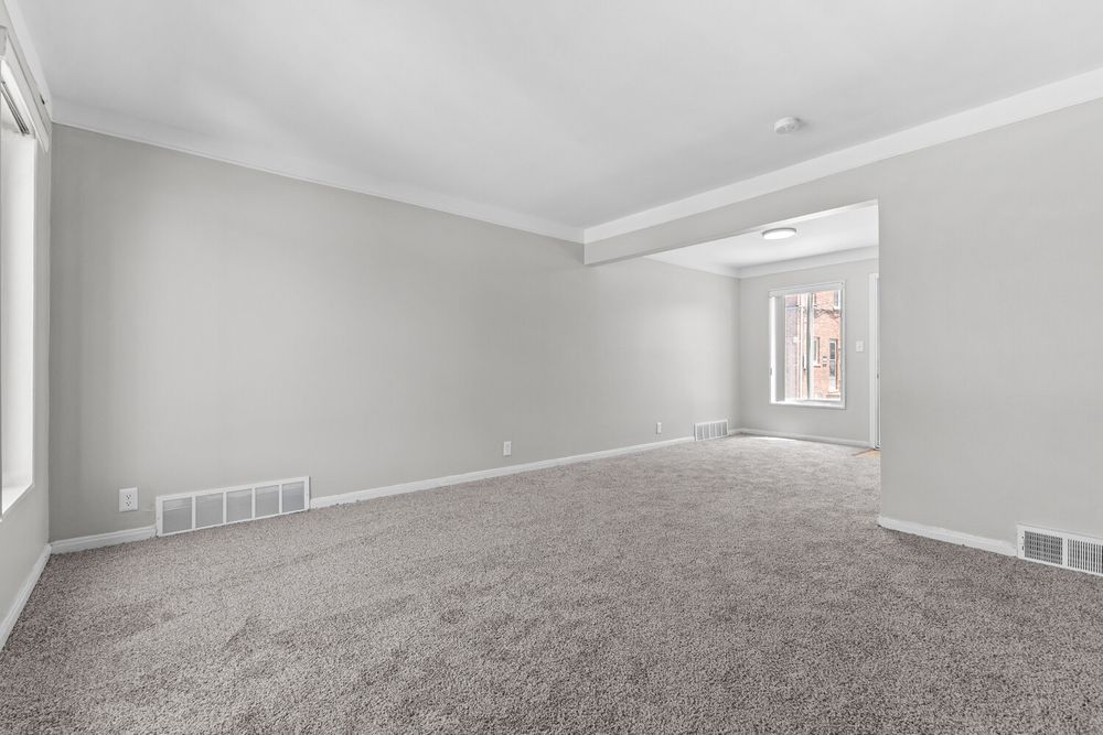 Spacious empty living room with gray carpet, crown molding, neutral walls, and a bright window passage.
