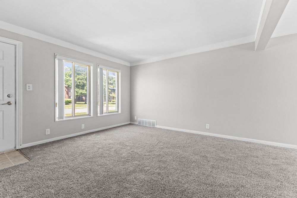 Bright empty living room with gray carpet, white walls, crown molding, and large windows.