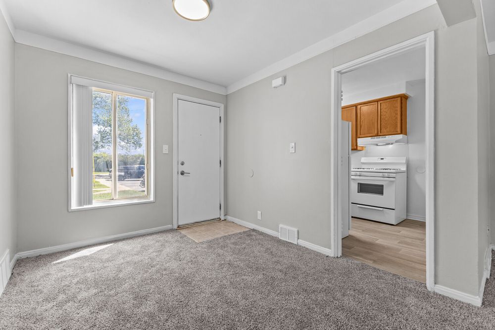 Bright dining area with gray carpet, crown molding, and a view into a kitchen with wood cabinets and white stove.