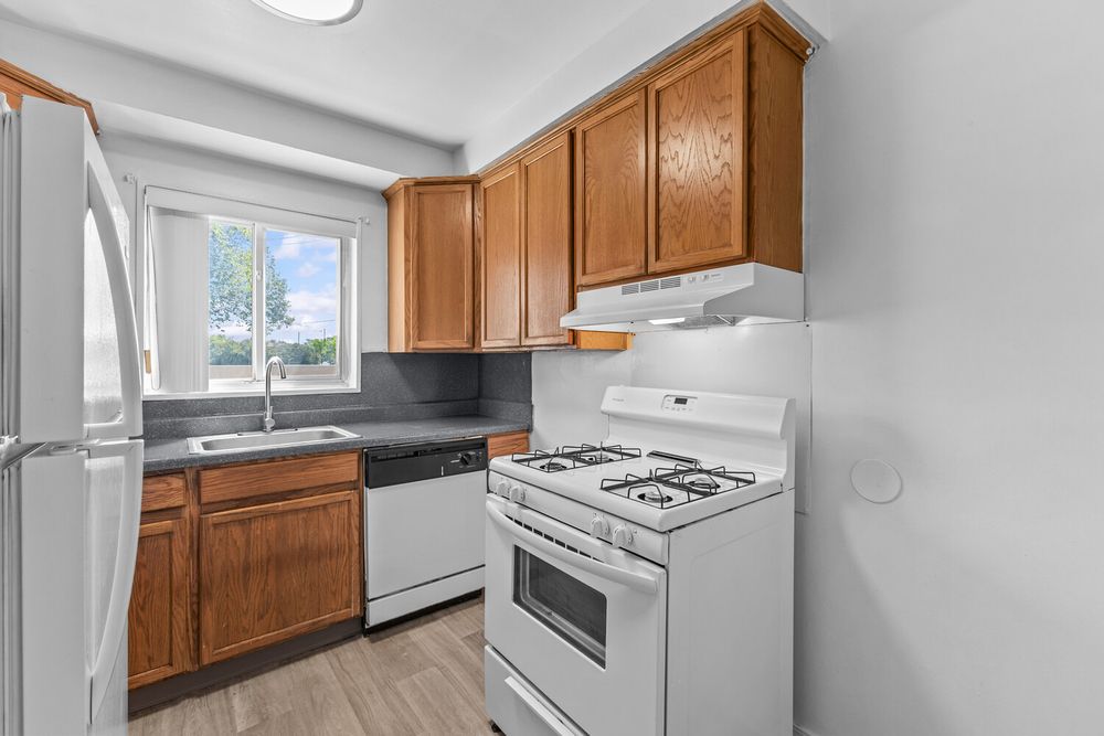 Kitchen with oak cabinets, white gas range, white dishwasher, dark countertops, and a window above the sink.