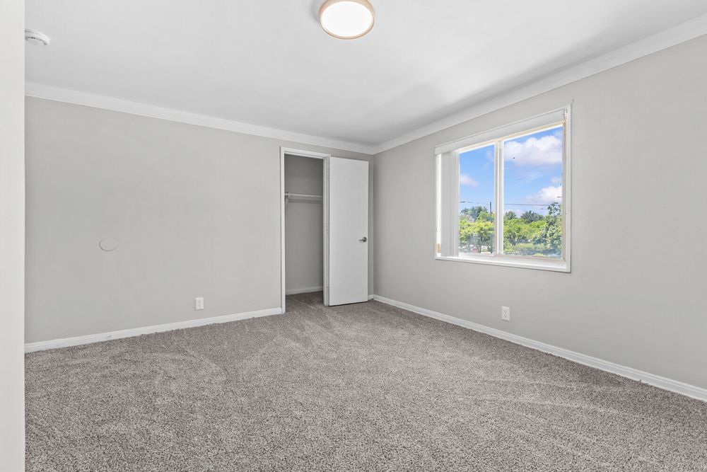 Empty bedroom with gray carpet, white walls, crown molding, open closet, and a bright window with outdoor views.