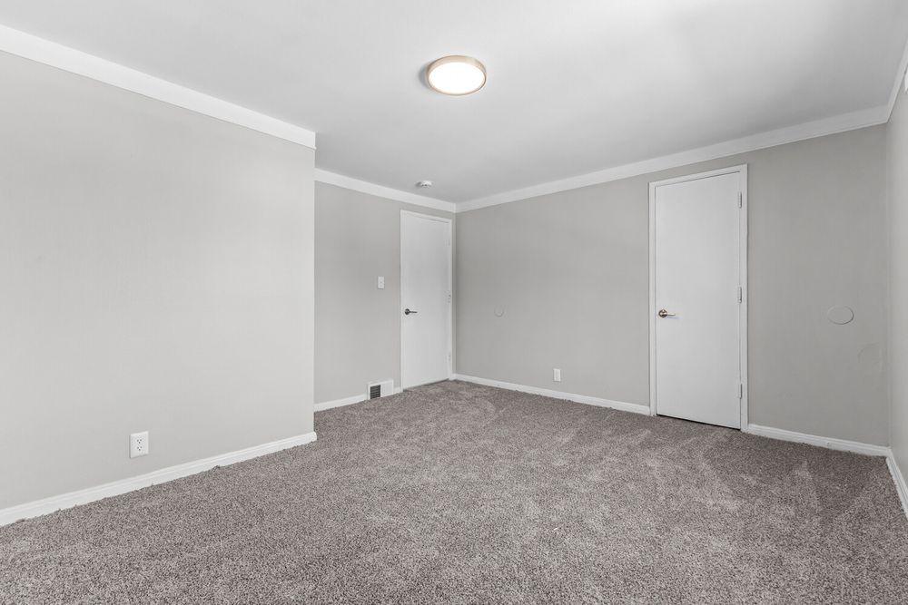 Spacious empty bedroom with gray carpet, crown molding, two white doors, and neutral gray walls with natural lighting.