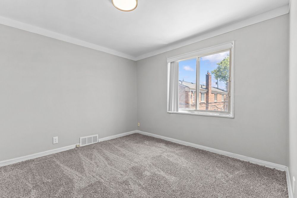 Bright bedroom with gray carpet, crown molding, white walls, and a window overlooking a brick building.