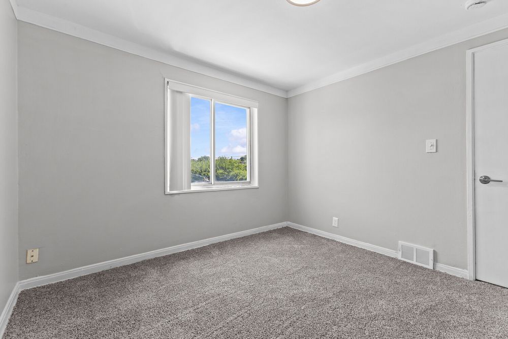 Empty bedroom with gray carpet, white walls, crown molding, and a window with natural light.