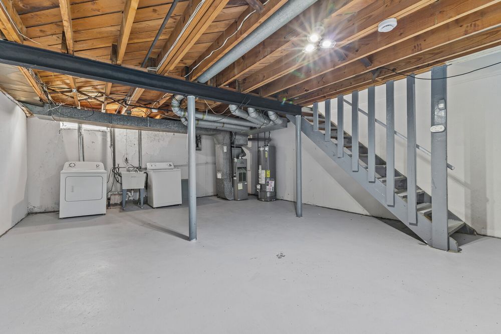 Unfinished basement with exposed wood ceiling, painted concrete floor, laundry area, and metal staircase.