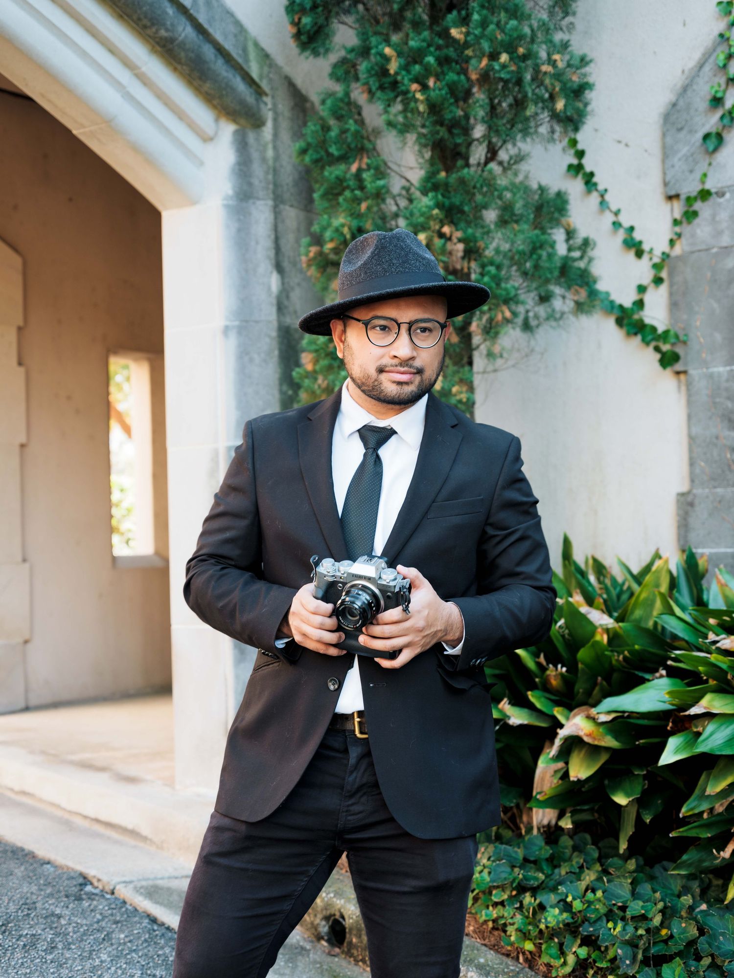 A stylish photographer in a black suit and hat holds a vintage camera outdoors near ivy-covered stone walls.