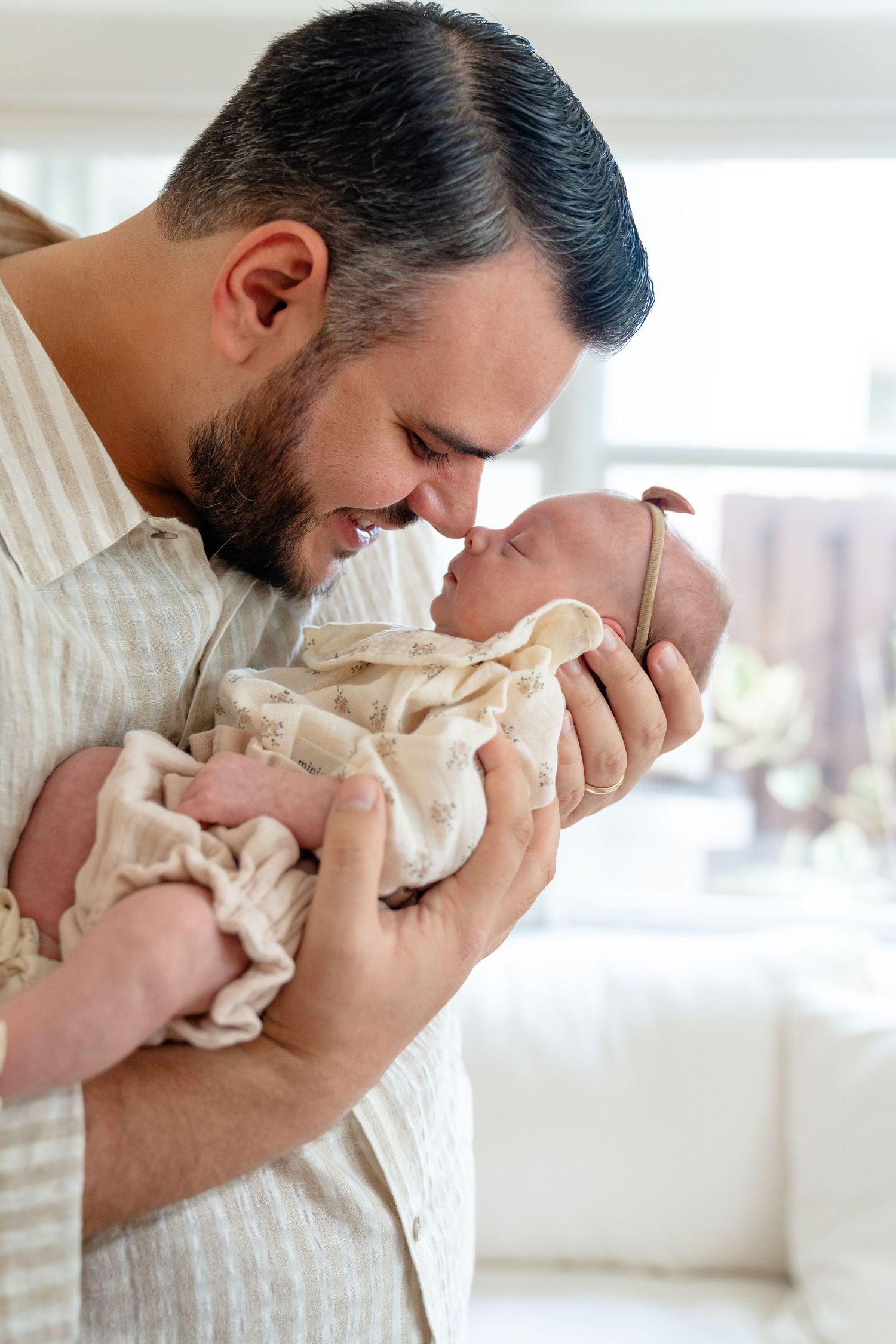 A bearded father tenderly holds his newborn baby, touching noses in a loving moment indoors.