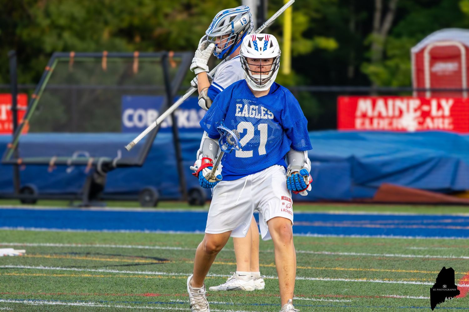 Two lacrosse players in blue and white uniforms compete during an outdoor game, one player shooting while the other defends.