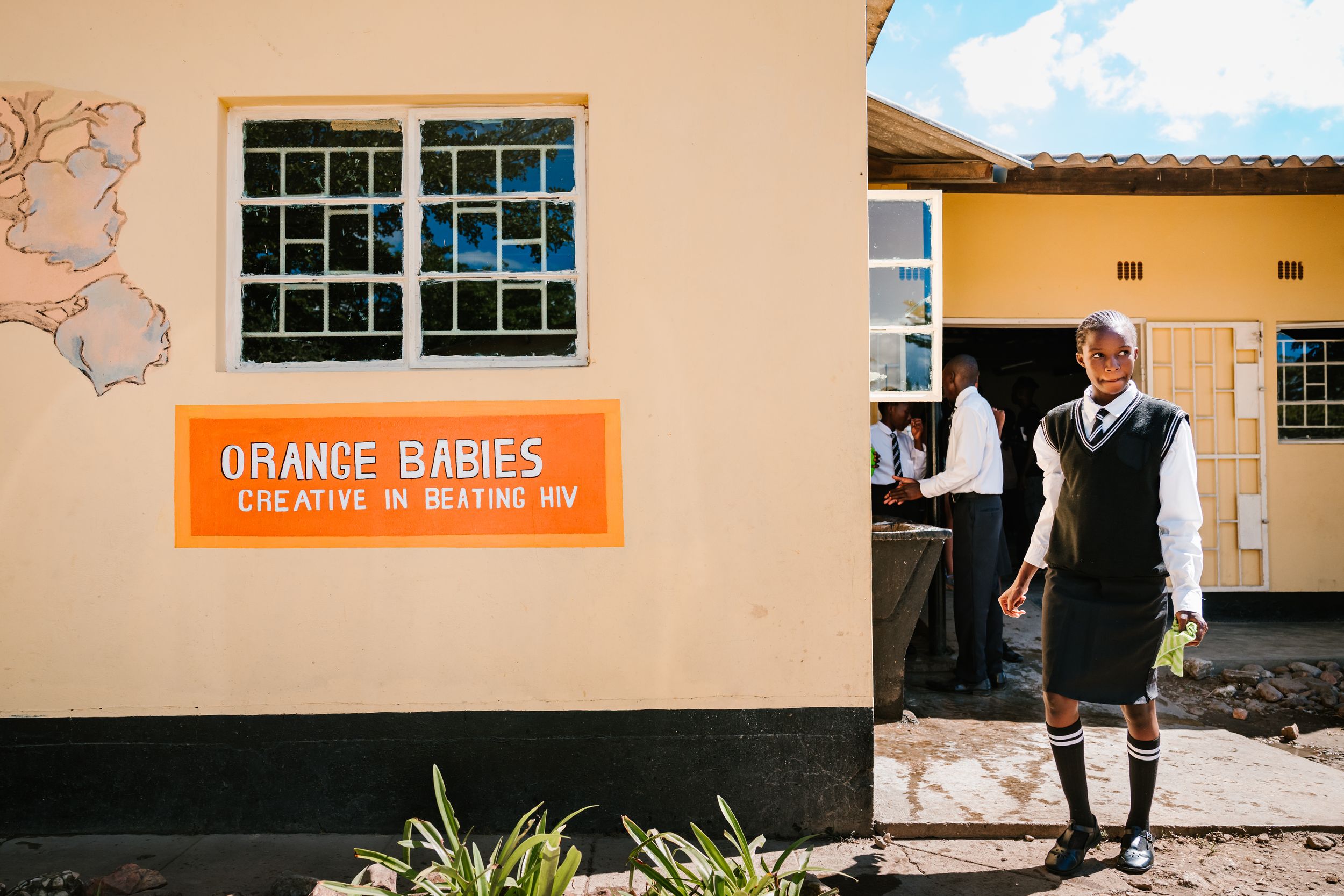 Student stands outside Orange Babies HIV awareness center in Africa, wearing school uniform.