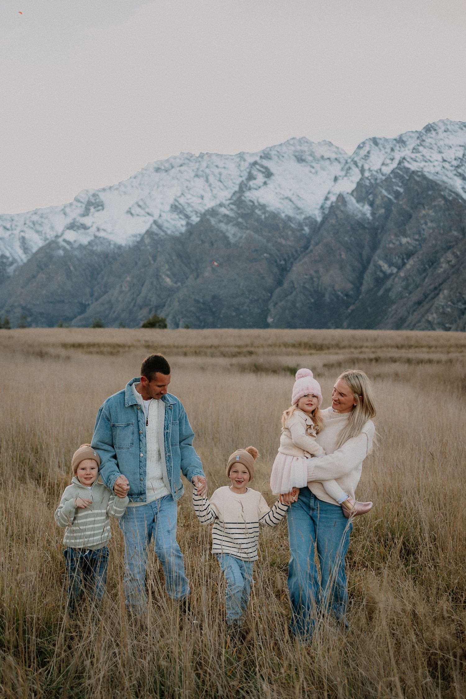Family of five posing in golden grass field with snow-capped mountains backdrop, parents holding toddlers with young child in center.