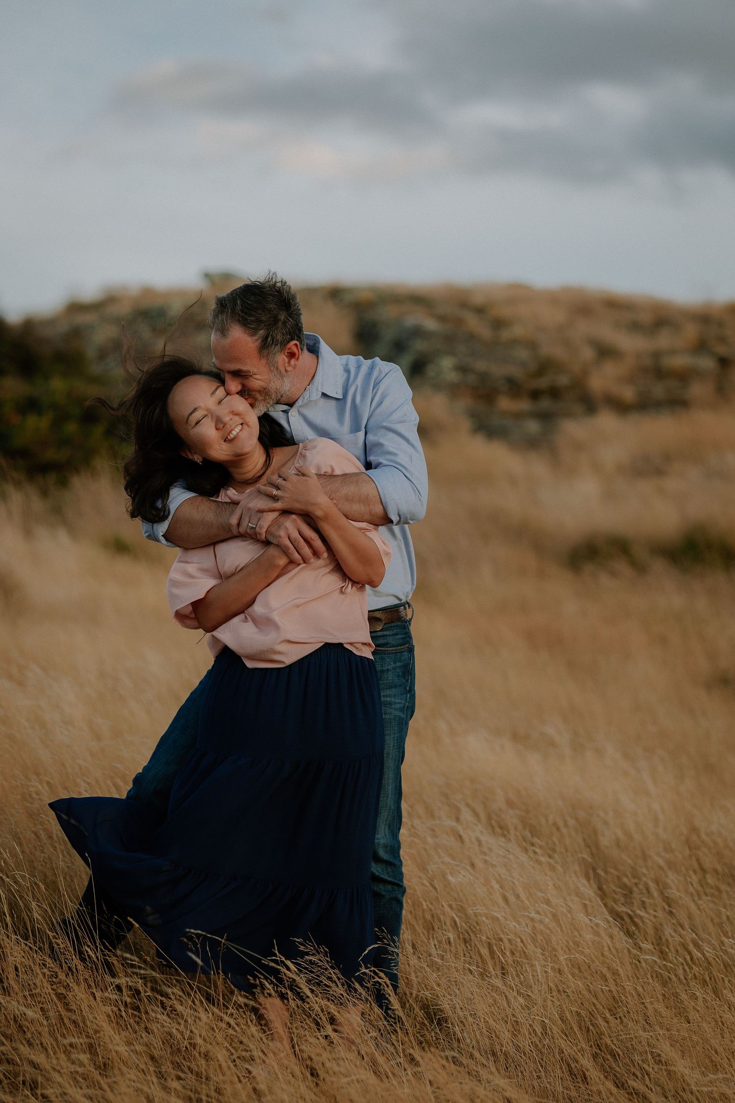 Couple embracing romantically in golden grass field, man kissing woman's cheek as she smiles, wearing pink top and navy skirt.