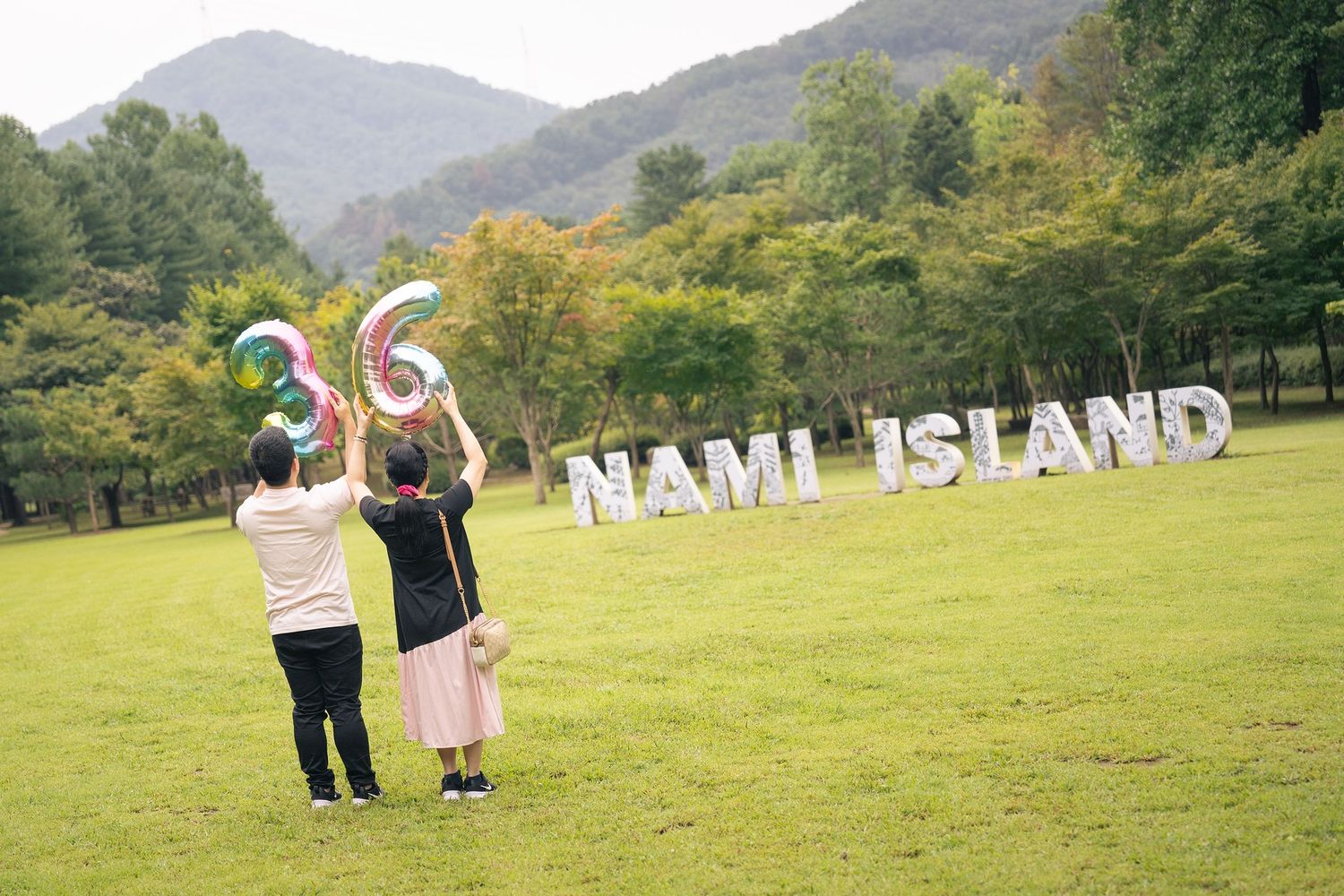 Two visitors hold number balloons at Nami Island, South Korea, posing in front of large silver letter signs on green lawn.