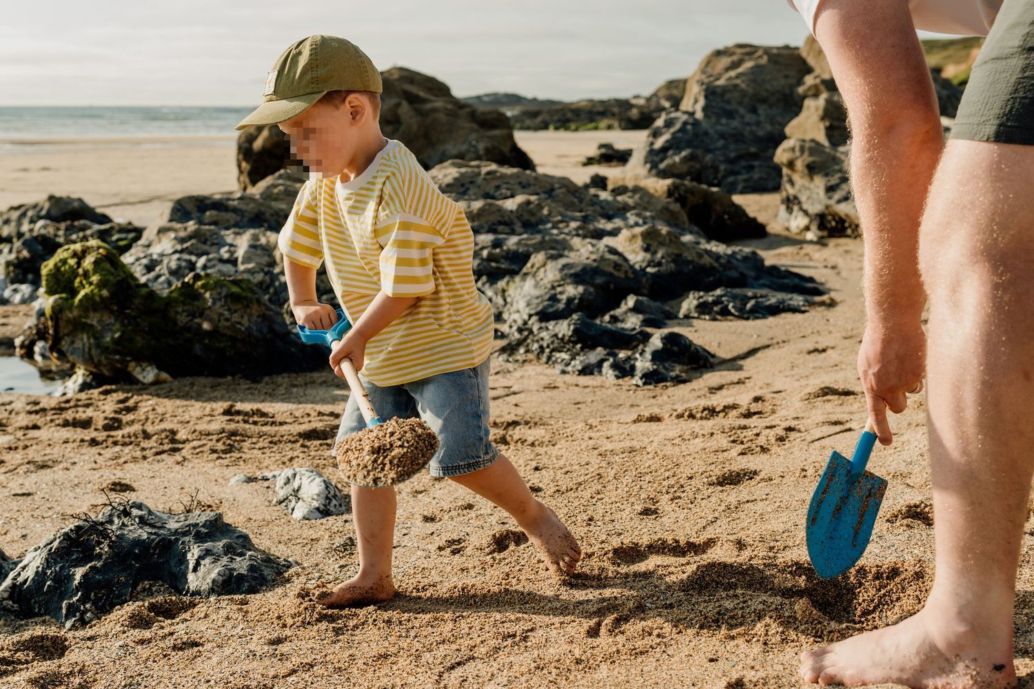 A young boy in a yellow striped shirt and cap plays with a blue shovel on a sandy beach near rocky shores.