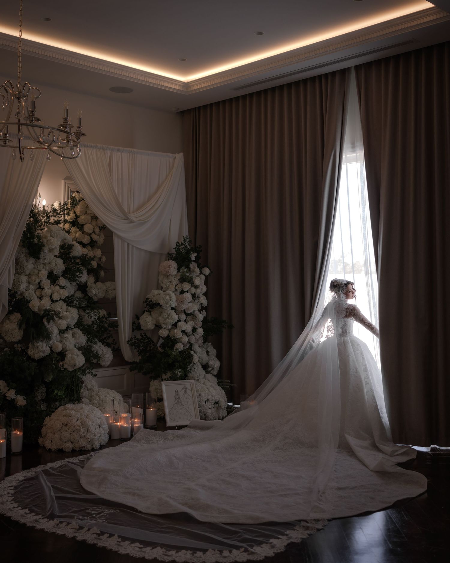 Bride in ball gown with long train standing in dimly lit room with white florals.