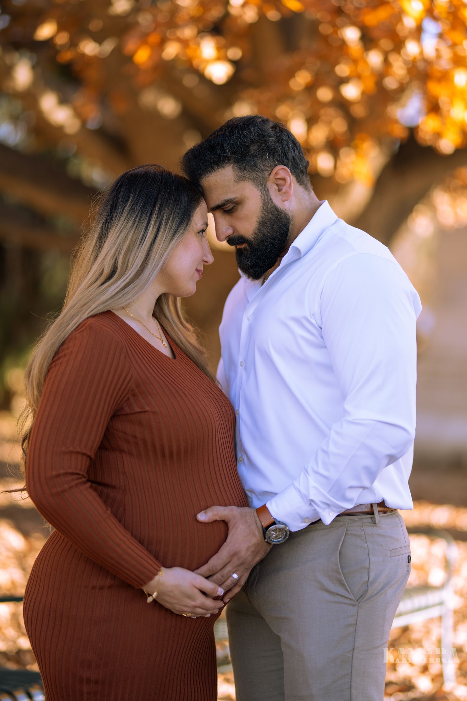 Pregnant couple embracing outdoors in autumn, woman in rust dress, man in white shirt, touching baby bump.