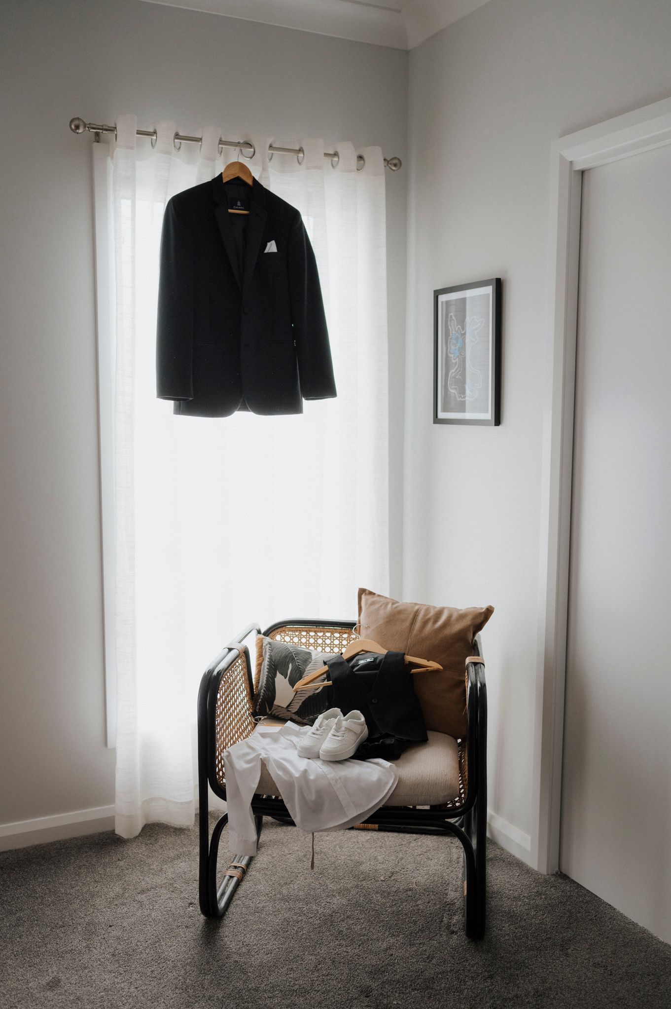 Black blazer hanging on curtain rod above a rattan chair with white sneakers and tan pillow in a minimalist room.