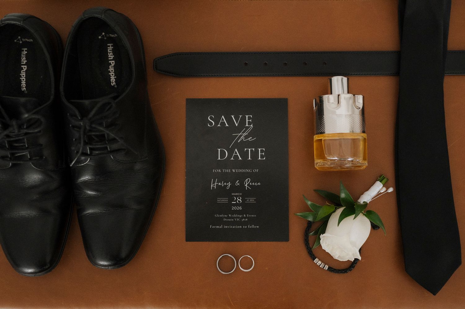 Black dress shoes, belt, tie, save the date card, wedding rings, boutonniere, and cologne arranged on a brown leather surface.