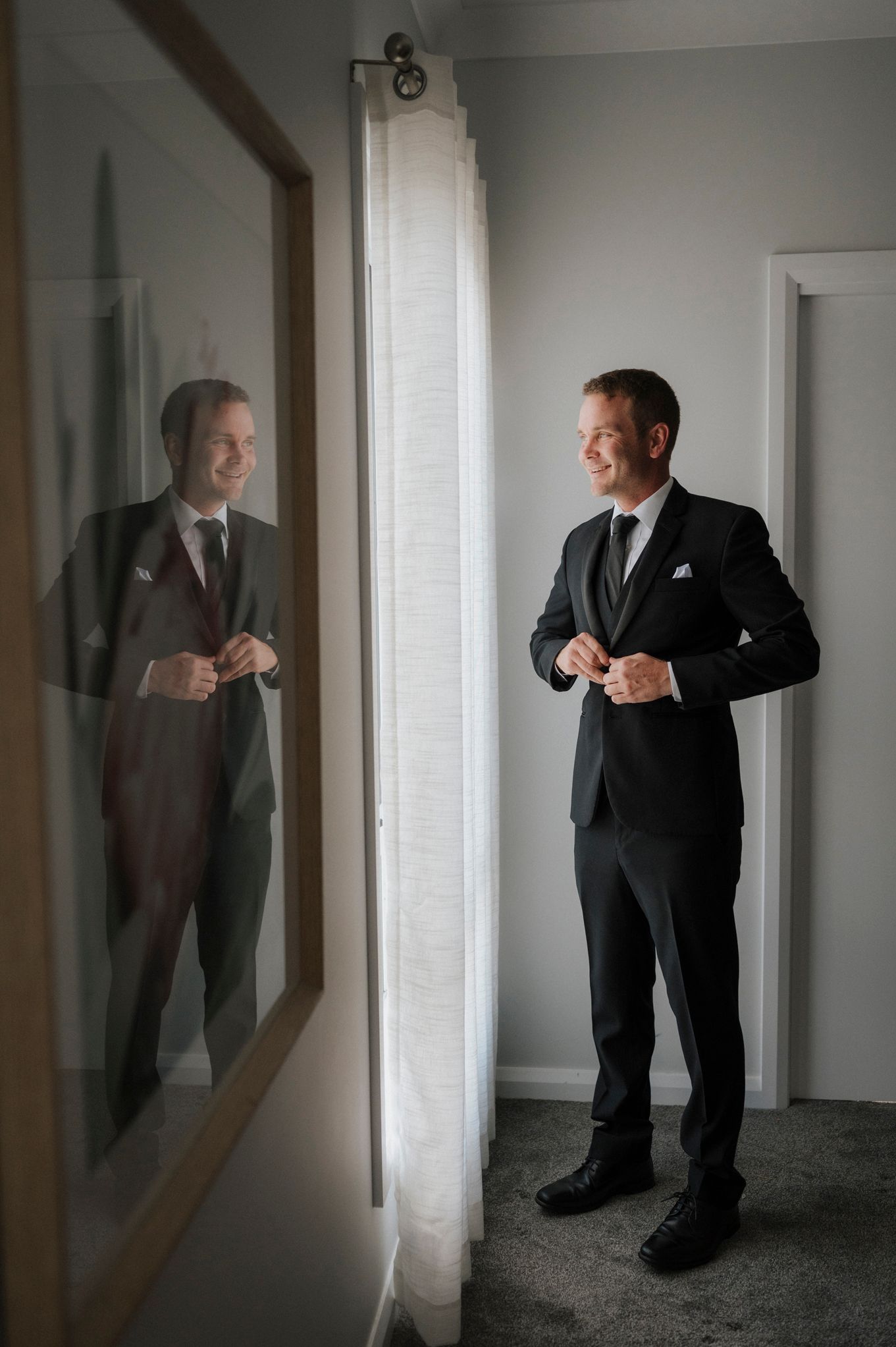 A smiling groom in a black suit buttons his jacket while standing near a mirror reflecting his image in a bright room.