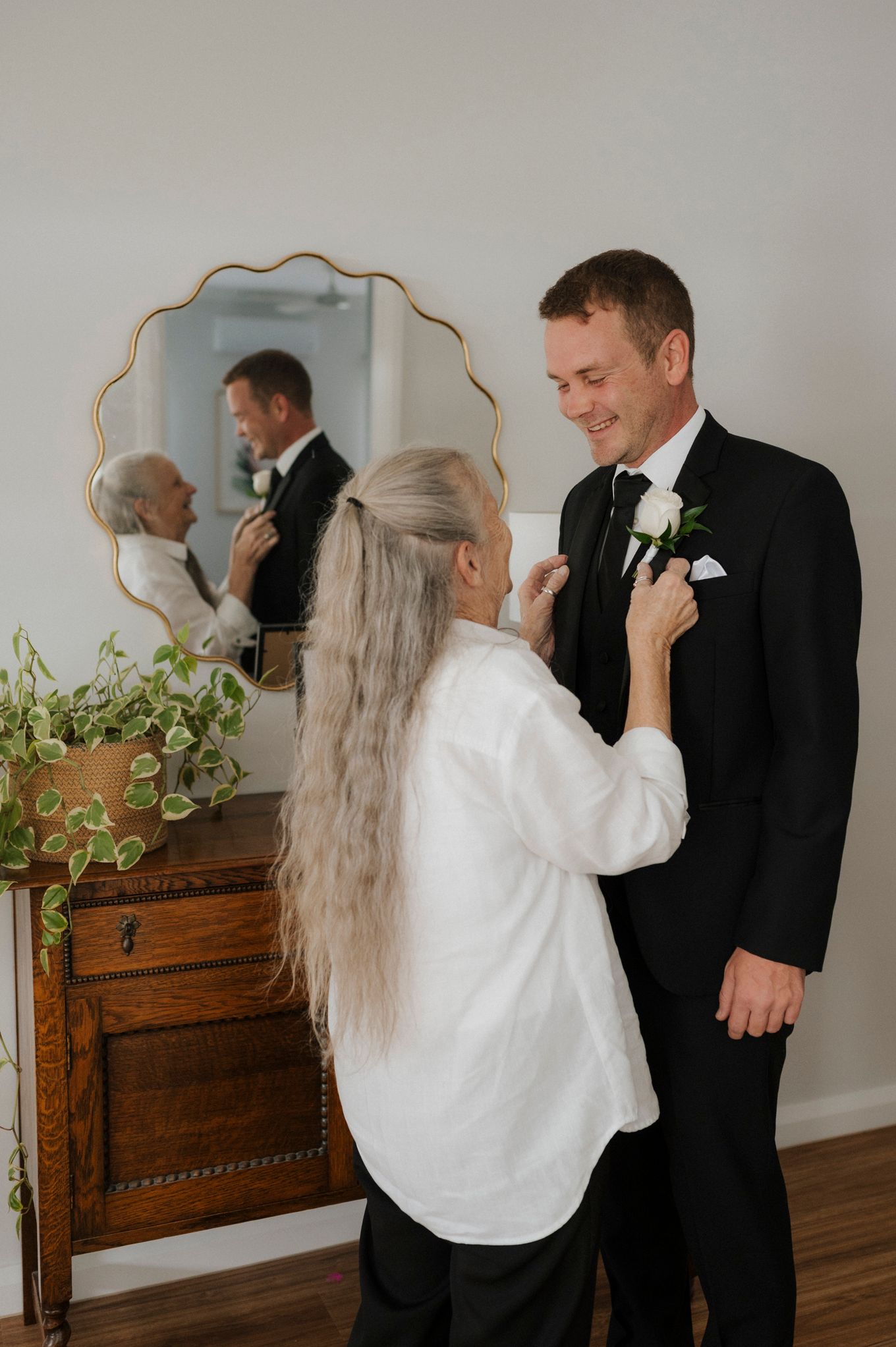 A woman pins a white boutonniere on a groom in a black tuxedo, reflected in a gold mirror above a wooden dresser.