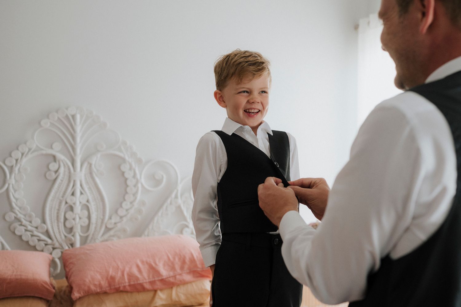 A young boy in a black vest and white shirt smiles while an adult adjusts his outfit before a formal event.